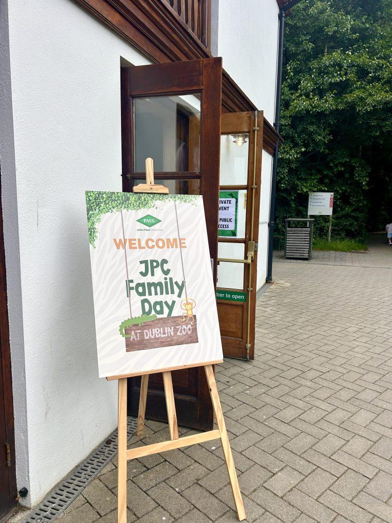 A wooden easel holding a welcome sign that reads "Welcome to JPC Family Day at Dublin Zoo" with cartoon animal illustrations, standing outside an open doorway.