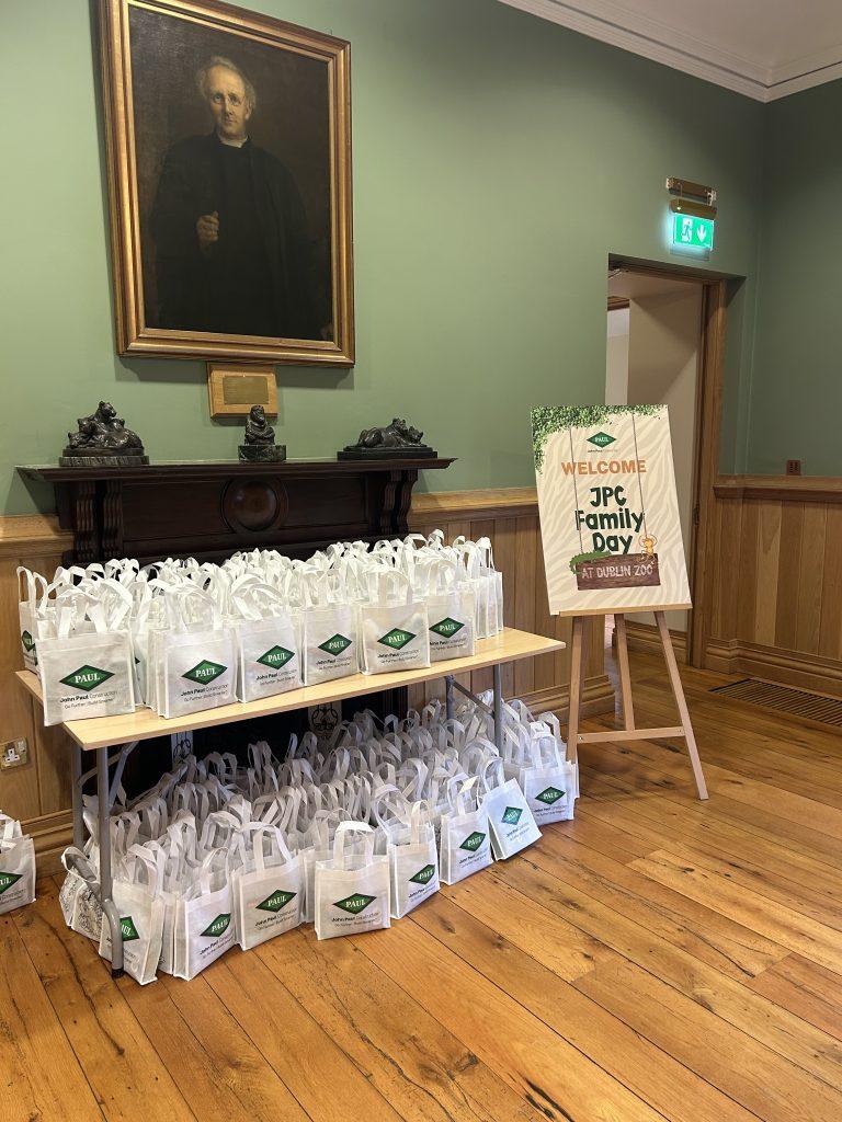 Dozens of white John Paul Construction branded gift bags arranged on and under a table indoors next to a "JPC Family Day" welcome sign.