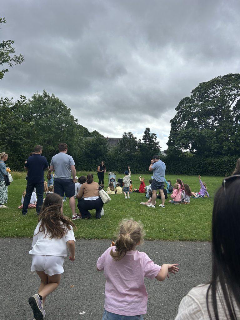 Parents and children gathered on a green lawn watching an outdoor magic show performance under an overcast sky at the Dublin Zoo family event.