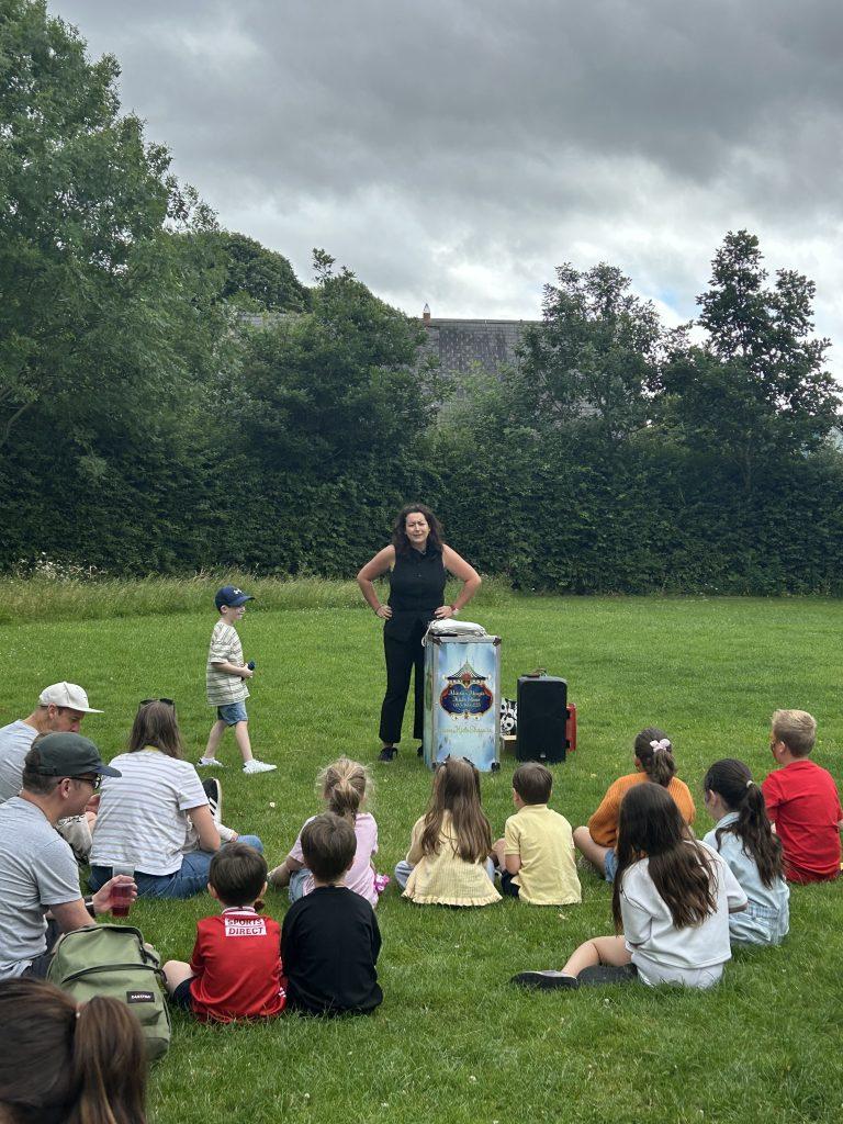 Children sitting on the grass watching a female magician perform next to a decorated prop box during the John Paul Construction family day.