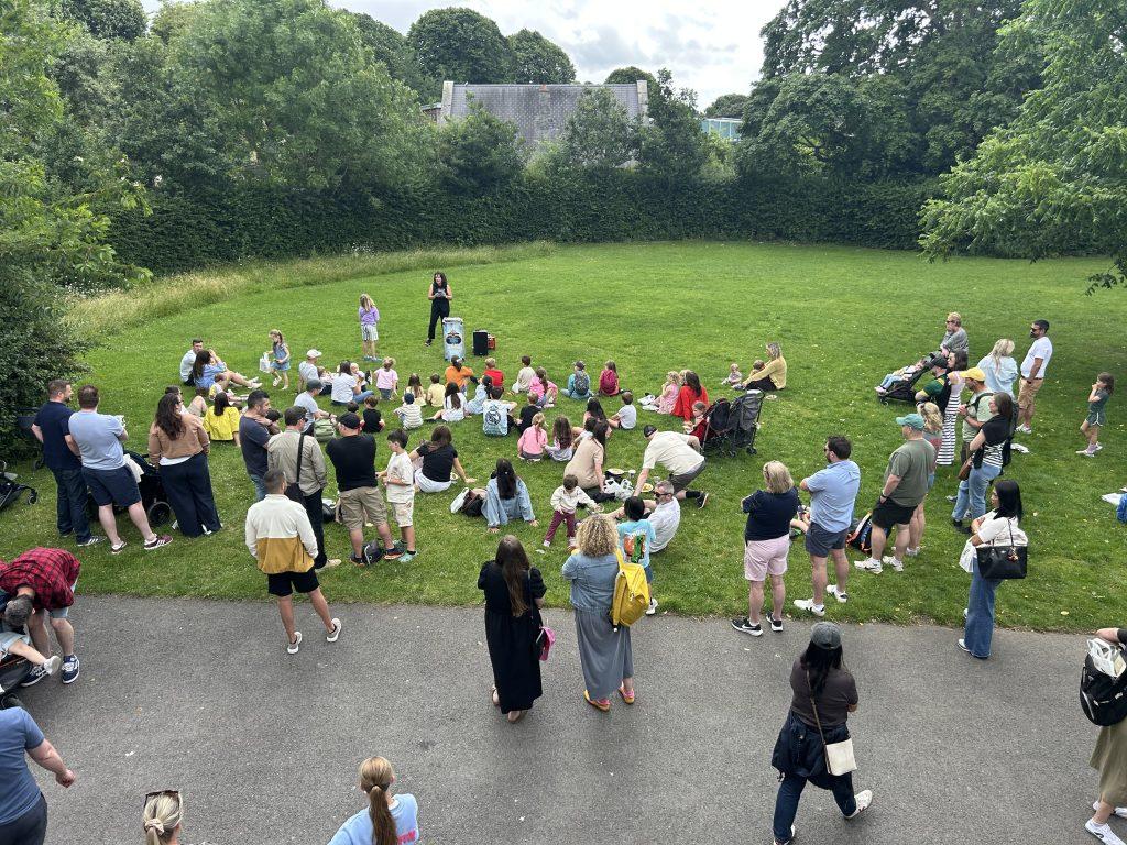 High-angle wide shot of a large group of John Paul Construction employees and their families gathered on a spacious green lawn enjoying an outdoor event at Dublin Zoo, watching the female magicians show. 
